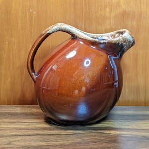 May include: A brown glazed ceramic pitcher with a rounded body and a handle. The pitcher has a light brown glaze with a mottled effect.