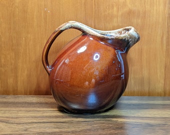May include: A brown glazed ceramic pitcher with a rounded body and a handle. The pitcher has a light brown glaze with a mottled effect.