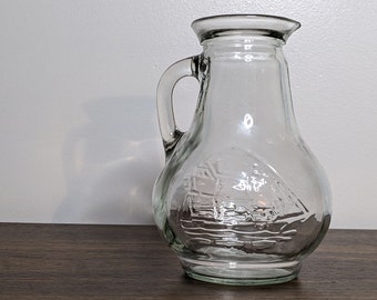 May include: A clear glass pitcher with a handle and a textured design of a sailboat on the front.