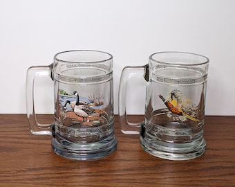 May include: Two clear glass beer mugs with handles. One mug features a painted scene of geese near a lake, the other a pheasant. Both mugs have a decorative band around the top and bottom.