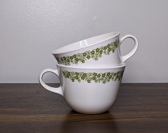 May include: Two white ceramic coffee cups stacked on top of each other. Each cup has a curved handle and a green floral pattern around the rim. The cups are sitting on a dark wooden surface.