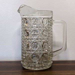 May include: A clear glass pitcher with a diamond pattern. The pitcher has a handle and a wide mouth.