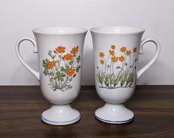 May include: Two white ceramic mugs with footed bases and floral designs. The mugs have orange and green floral patterns and blue trim around the base.