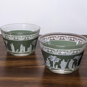 May include: Two clear glass bowls with a dark green band featuring white classical figures and decorative patterns. The bowls are on a dark wooden surface.