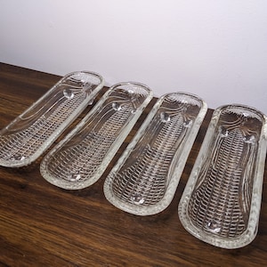 May include: Four clear glass corn on the cob serving dishes. The dishes are designed to hold corn on the cob and have a textured surface that resembles corn kernels.