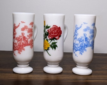 May include: Three white ceramic mugs with different floral and figurative designs. The mugs have a white background with red, blue, and yellow floral designs. The mugs have a handle on the side and a rounded base.