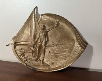 May include: A gold-toned metal ashtray with a raised design of a sailor standing on a boat with a sail. The boat is on a body of water with other boats in the background.