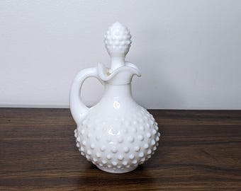 May include: A white hobnail glass cruet with a handle and a stopper. The cruet has a bulbous body covered in small, raised bumps. The stopper is also white and has a textured, rounded top. The cruet is sitting on a dark wooden surface.