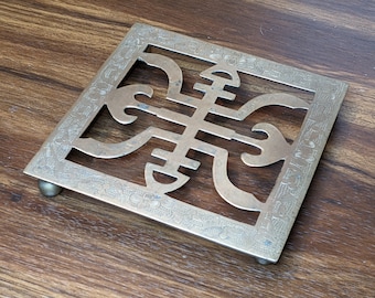 May include: A square brass trivet with an intricate design. The trivet has a raised, openwork pattern in the center and a decorative border around the edges.