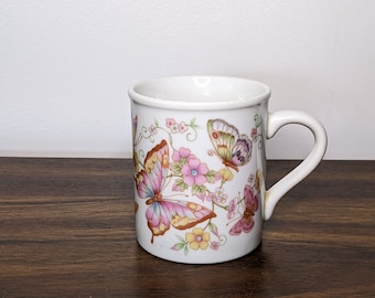 May include: White ceramic mug with a butterfly and floral design. The butterflies are pink, yellow, and green. The flowers are pink, yellow, and white.