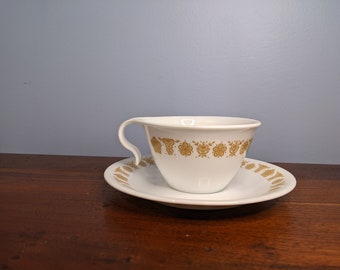 May include: A white teacup and saucer with a gold floral pattern. The cup has a unique handle that curves around the side.