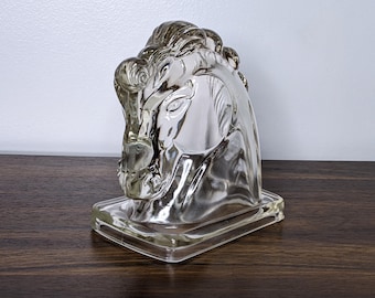 May include: A clear glass horse head sculpture. The horse head is detailed with a flowing mane and sits on a rectangular base. The sculpture is displayed on a dark wood surface.