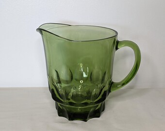 May include: A green glass pitcher with a handle and a spout. The pitcher has a textured design on the lower portion and a smooth upper portion. The handle is curved and attached to the side of the pitcher. The pitcher is likely used for serving beverages.