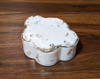 May include: A white porcelain trinket box with gold accents and a floral design. The box has a rounded, four-lobed shape.