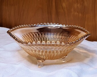 May include: An amber-colored, footed glass bowl with a beaded rim. The bowl has a diamond pattern on the sides and three small feet. The bowl is likely vintage and suitable for serving small portions.