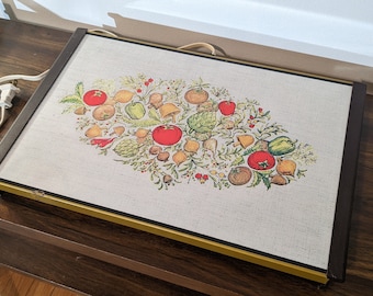 May include: Vintage electric warming tray with a beige linen-like surface. The tray features a colorful illustration of various vegetables, including tomatoes, peppers, and mushrooms. The tray has a brown frame with a gold trim and a power cord.