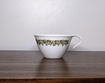 May include: White teacup with a green floral pattern around the rim. The cup has a curved handle and sits on a dark wooden surface. The background is a plain white wall.