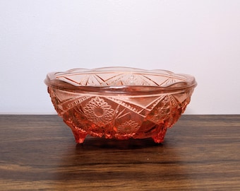 May include: A pink glass bowl with a geometric pattern and three legs. The bowl is on a wooden surface.