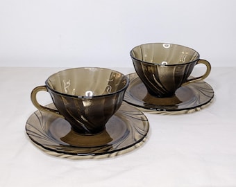 May include: Two vintage brown glass teacups and saucers. The cups and saucers have a spiral design. The cups have handles. The saucers have a ridged edge. The set is on a white surface.