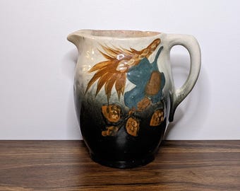 May include: A ceramic pitcher with a handle and spout. The pitcher is painted with a floral design in shades of brown, blue, and green against a cream and black background. The pitcher is sitting on a wooden surface.