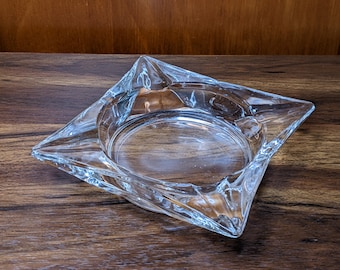 May include: A clear glass ashtray with a square shape and a slightly raised center. The ashtray has a faceted design and is sitting on a wooden surface.