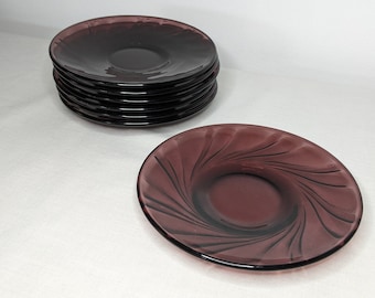 May include: A stack of six purple glass saucers with a single saucer with a swirl design in the foreground.