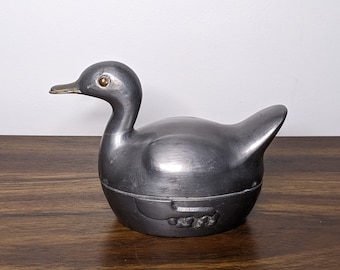 May include: A silver metal duck figurine with a hinged lid. The duck has a detailed design and a gold eye.