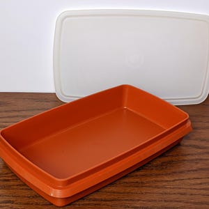 May include: An orange rectangular food storage container with a white lid. The container is made of plastic and has a slightly raised rim. The lid is translucent and rectangular. The container is on a wooden surface.