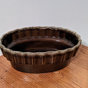 May include: A brown ceramic pie dish with a scalloped edge. The dish has a dark brown interior and a lighter brown exterior with vertical ridges. The dish sits on a wooden surface.