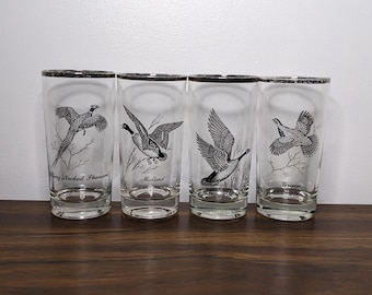 May include: Set of four clear glass drinking glasses with black and white illustrations of different birds. The glasses feature the names "Ring Necked Pheasant", "Mallard", "Canada Goose", and "Grouse".