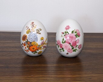 May include: Two white ceramic eggs with floral designs. One egg has a pink rose pattern, and the other has a multi-colored floral pattern.