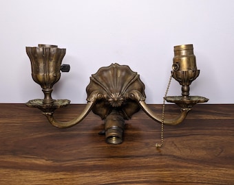 May include: Antique brass wall sconce with two light fixtures. The sconce features a central decorative element with a shell-like design and curved arms. Each light fixture has a decorative base and a bulb socket. A pull chain hangs from one socket.