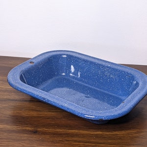 May include: A blue enamelware rectangular baking dish with white speckles. The dish has rounded corners and small holes on the sides. This vintage-style dish is suitable for baking and serving.