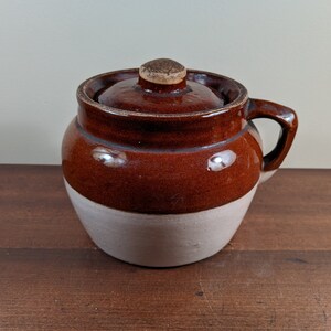 May include: A brown and white glazed ceramic crock with a lid and a handle. The crock is a traditional style with a rounded body and a wide mouth.