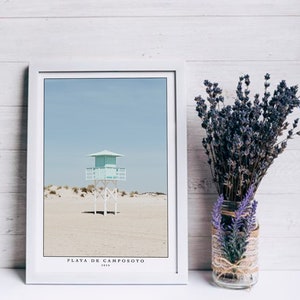 Puede incluir: Una fotografía enmarcada de una caseta de salvavidas blanca con un techo azul claro en una playa de arena. El cielo es azul claro y el texto "PLAYA DE CAMPOSOTO 2010" está en la parte inferior de la imagen.