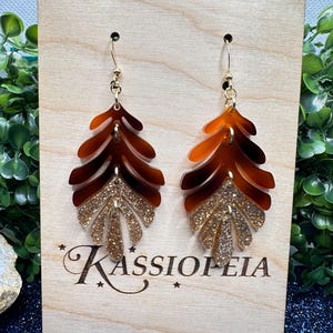 May include: A pair of brown and gold glitter leaf-shaped earrings with gold hooks. The earrings are displayed on a wooden board with the text "Kassiopeia" engraved on it.