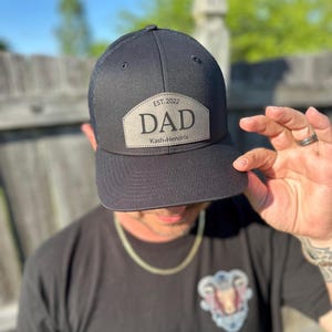 May include: Black trucker hat with a curved brim and mesh back. The hat features a light gray leather patch with the word "DAD" in bold letters, the year "EST. 2022", and the name "Kash-Hendrix".