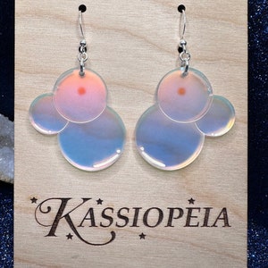 Peut inclure: Une paire de boucles d'oreilles irisées avec un design à trois bulles. Les boucles d'oreilles sont en argent et ont une finition irisée rose et bleue. Les boucles d'oreilles sont sur une carte d'affichage en bois avec le texte "KASSIOPEIA" imprimé dessus.