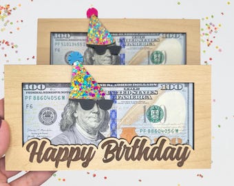 May include: A wooden frame with a $100 bill inside. The frame has a cutout for a birthday message and a party hat with sunglasses on top. The message reads "Happy Birthday".