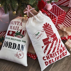 May include: Two off-white canvas drawstring bags for gifts. The left bag features red and green text reading SANTA MAIL, SPECIAL DELIVERY FOR JORDAN, and a North Pole stamp. The right bag shows a stack of red gifts and text reading CHRISTMAS DELIVERY.