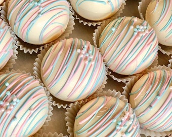 May include: A collection of white chocolate spheres, decorated with pink and pale blue stripes and small sprinkles. Each sphere sits in a white paper case. The arrangement suggests a dessert or confectionery item, possibly for a celebration.