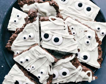 Könnte beinhalten: Ein schwarzer Teller mit dreieckigen Schokoladenstücken, die mit weißem Zuckerguss in bandagenartigem Muster und kleinen runden Zuckeraugen verziert sind. Der dunkle Schokoladenboden ist unter dem Zuckerguss und den Dekorationen sichtbar.