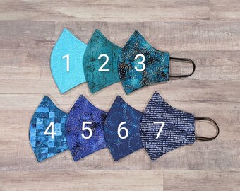 May include: Seven different fabric face masks in various shades of blue and green with gold and white accents. The masks are numbered 1 through 7.