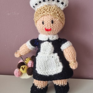 Könnte beinhalten: Eine gestrickte Puppe, die eine weiße Mütze mit schwarzem Besatz, ein schwarzes Kleid mit einer weißen Schürze und schwarze Schuhe trägt. Die Puppe hält einen Korb mit bunten Süßigkeiten.