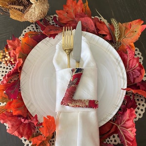 May include: A white napkin with a gold fork and knife is wrapped around a fabric letter 'L' and placed on a white plate. The plate is surrounded by a brown placemat with red and orange fall leaves.