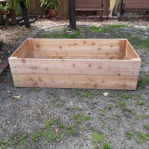 May include: A rectangular wooden raised garden bed made of untreated cedar planks. The bed is empty and sitting on a gravel surface.