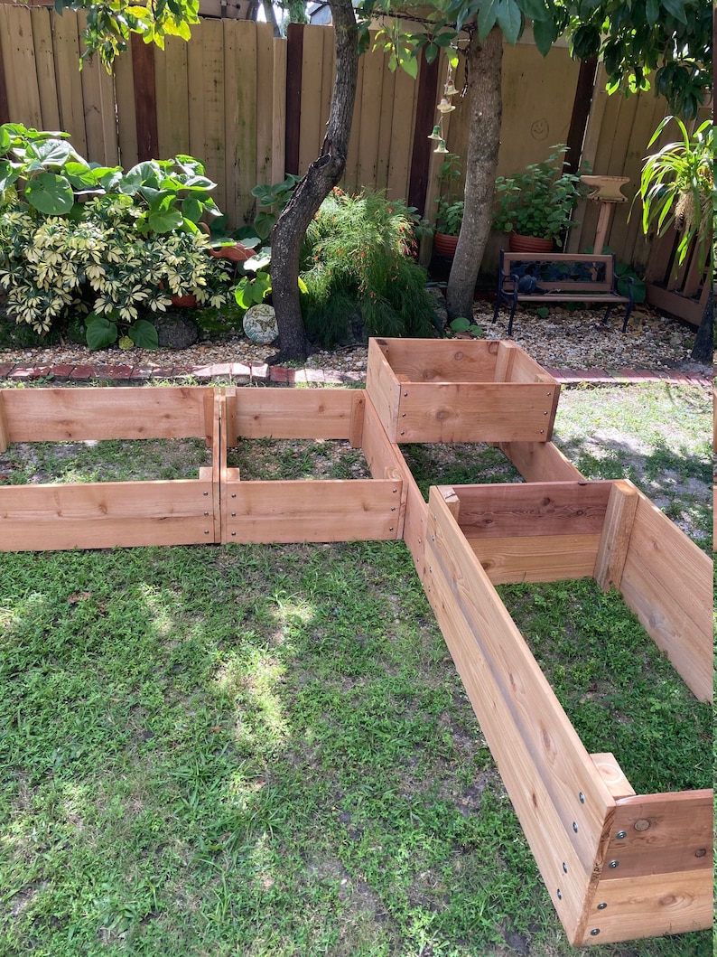 May include: Four wooden raised garden beds in a backyard setting. The beds are made of cedar wood and are arranged in a staggered pattern. The beds are surrounded by green grass.