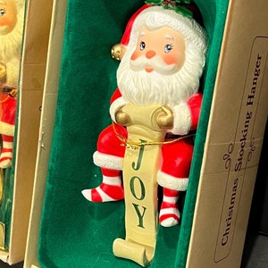 May include: A white porcelain Christmas stocking hanger featuring a Santa Claus figurine holding a scroll with the word "JOY" written on it. The hanger is packaged in a box with a green velvet lining and the words "Christmas Stocking Hanger" printed on the side.