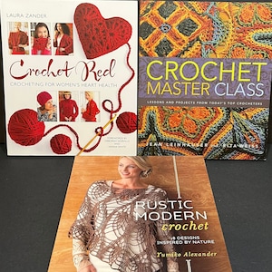May include: Three crochet books with colorful covers. The first book is titled "Crochet Red: Crocheting for Women's Heart Health" and features a red heart and yarn. The second book is titled "Crochet Master Class: Lessons and Projects from Today's Top Crocheters" and features a colorful crochet design. The third book is titled "Rustic Modern Crochet: 18 Designs Inspired by Nature" and features a woman wearing a brown crochet shawl.
