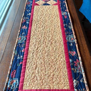May include: A quilted table runner with a central beige section featuring a repeating pattern. It is bordered by a dark blue fabric with a floral design and a red accent. The runner is placed on a wooden surface.